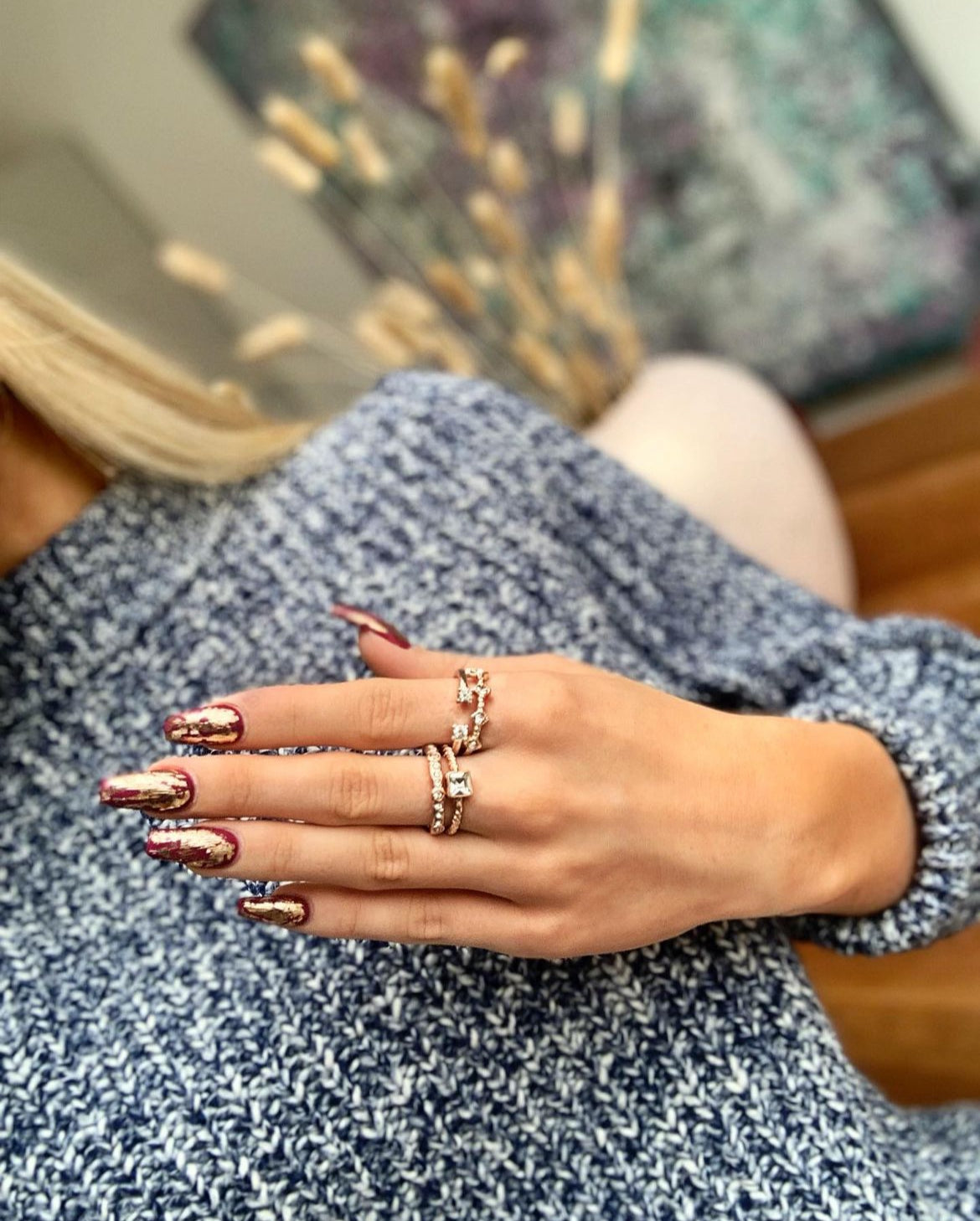 Tjej med blå tröja och blont hår visar sin hand med röda långa naglar med guldiga detaljer och tre ringar på fingrarna.